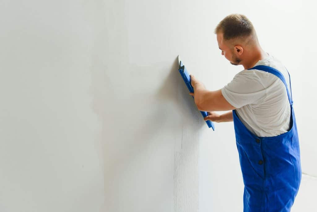 Ouvrier en bleu de travail appliquant du ciment gris sur un mur blanc à l'aide d'une truelle.