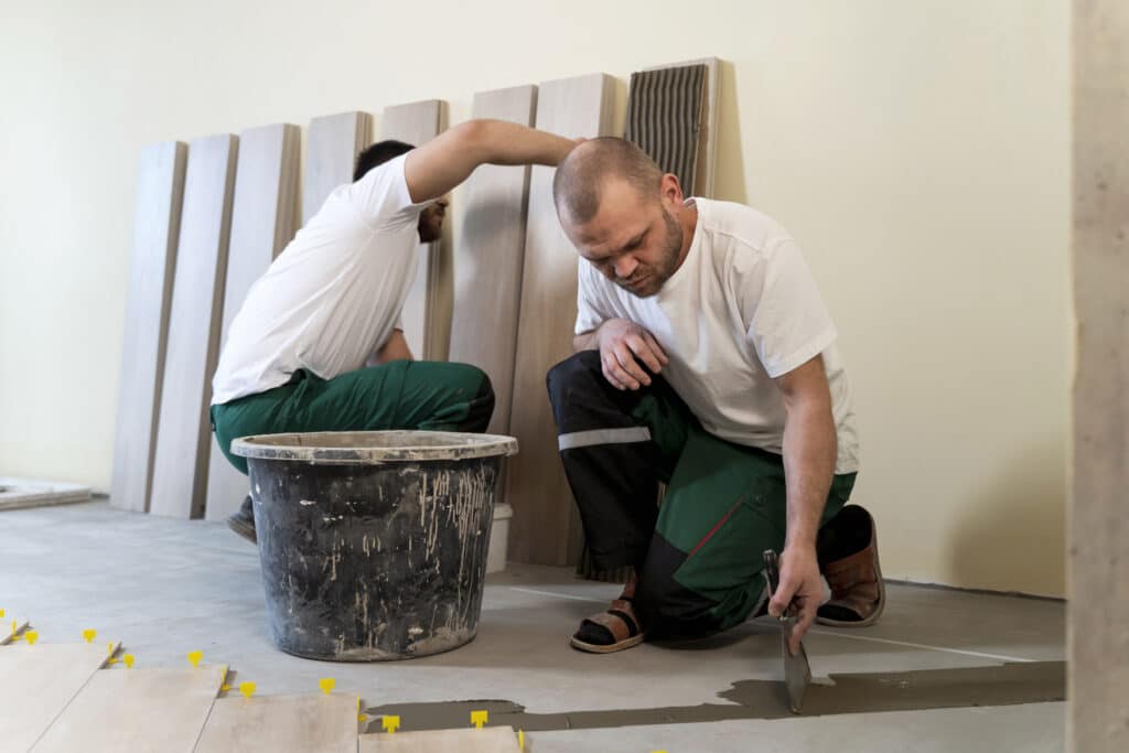 Deux carreleurs au travail, l'un reposant tandis que l'autre coupe des carreaux de céramique pour le sol.