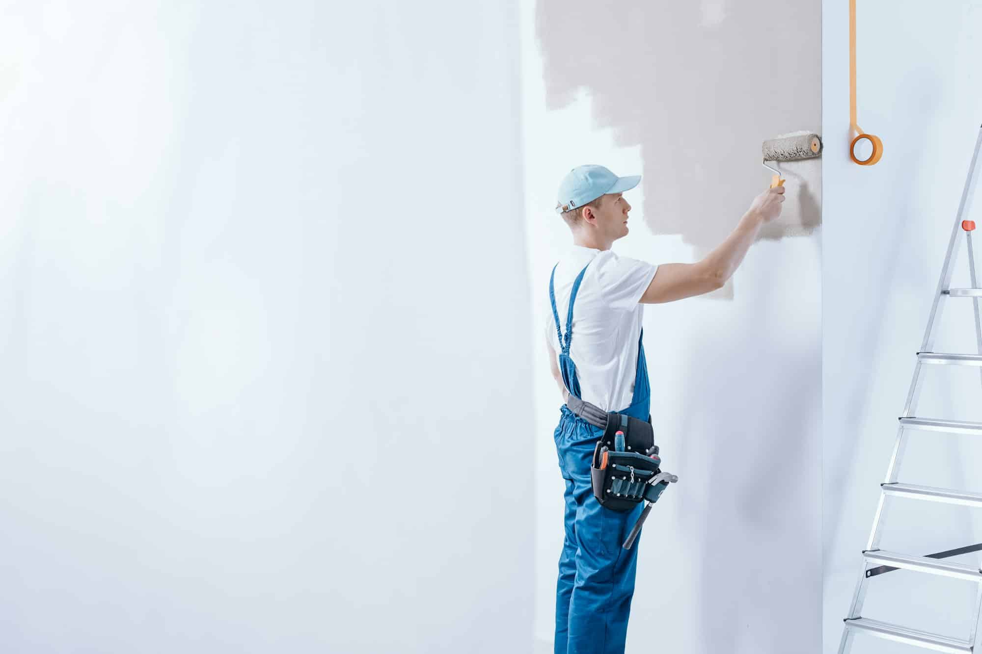 Peintre en combinaison bleue peignant un mur intérieur blanc à l'aide d'un rouleau de peinture.