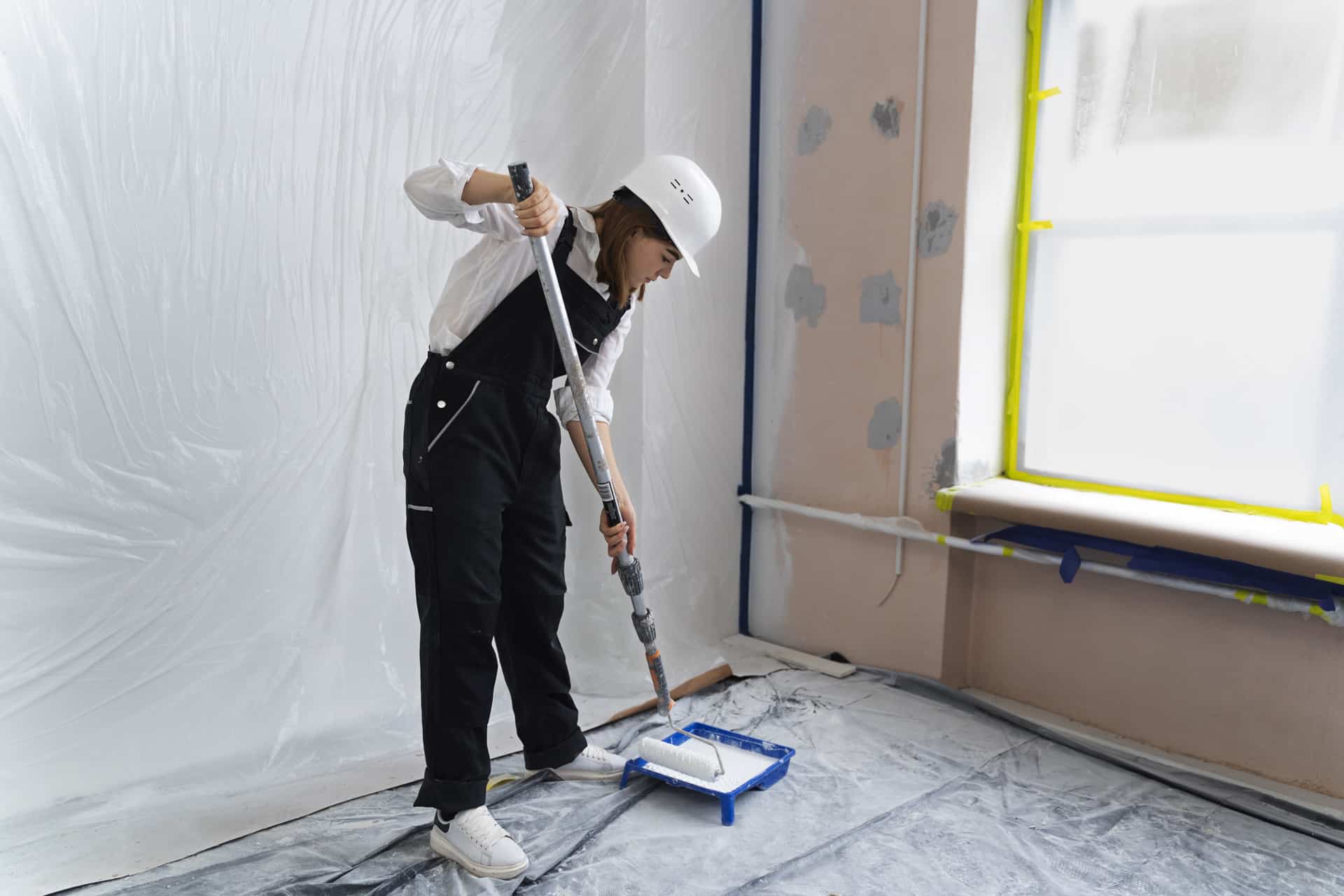 Femme en tenue de protection blanche peignant un mur d'une pièce recouverte de bâches en plastique.