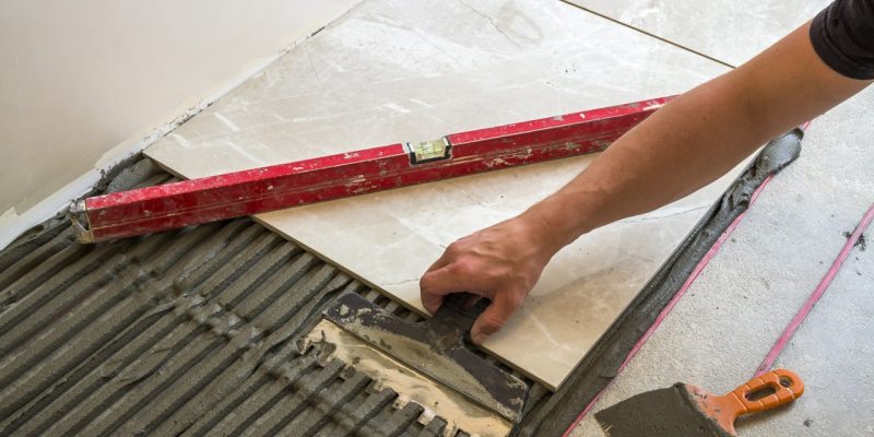 Main d'un travailleur posant des carreaux de céramique blancs sur un sol avec une truelle et du mortier.